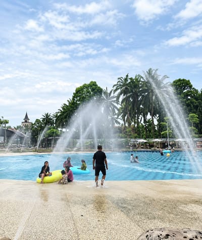 This image depicts a water park scene in what is identified as Eampat Kwalamencirim, Indonesia. The foreground shows a textured, light-colored surface with water splashing on it, indicating the edge of a pool. In the middle ground, a large blue swimming pool is visible, with multiple water jets creating spray and mist. Several people are in and around the pool.
In the immediate foreground of the pool's edge, a Black man wearing black shorts and a black t-shirt is walking away from the camera, with his feet visible in the splashing water. To his left, a group of people are near the edge of the pool. A woman in a black top and dark pants is sitting on a yellow inflatable ring. Beside her, a child is sitting on the ground. Further to the left, another woman in a pink top and dark pants is also sitting near the water. A young child is sitting on a green inflatable ring.
In the mid-ground of the pool, several individuals are swimming or floating. Two men are visible further back in the water. Another person is on a yellow inflatable ring.
The background features lush green trees and palm trees, characteristic of a tropical environment. A dark, tower-like structure with a pointed roof, resembling a castle or a distinctive building, is visible on the left side of the background, nestled amongst the trees. Umbrellas and seating areas are also visible in the distance near the pool. The sky above is bright blue with scattered white clouds. The overall impression is a lively recreational setting on a sunny day.