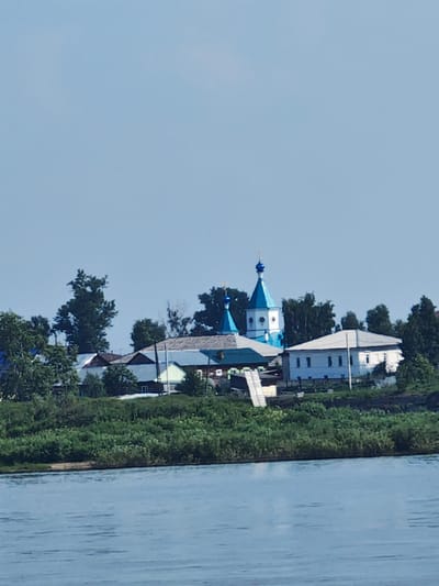 A wide shot captures a serene summer day on the Yenisei River in Yeniseysk, Russia. The calm, blue water fills the foreground, reflecting the clear sky above. The middle ground is dominated by a lush green embankment, thick with trees and vegetation, creating a vibrant natural barrier. Perched on this embankment is a small cluster of buildings, including a prominent white church with bright blue domes and spire. The church stands out against the backdrop of the sky and surrounding trees. Adjacent to the church are other residential or administrative buildings, also featuring light-colored walls and varied rooflines. The scene exudes a sense of peace and rural tranquility, typical of a riverside town in the Siberian region. There are no people visible in the image.