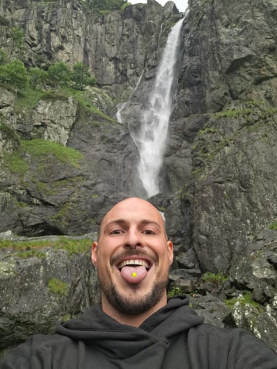 A man with a shaved head, a septum piercing, and a tongue piercing with a yellow stud is taking a selfie. He is wearing a black hooded sweatshirt. Behind him, a waterfall cascades down a rocky cliff face. The surrounding environment is mountainous and covered in green vegetation. The location is the waterfall in Tazha, Bulgaria.