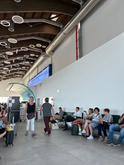 The image depicts the interior of a modern airport terminal in Lamezia Terme, Italy, likely the departure lounge. A woman in a black tank top and white pants walks towards the right, her back mostly to the camera. Another person in a gray t-shirt and maroon pants walks in the same direction, ahead of her. In the background, a group of people are seated on a bench, waiting. One man is looking at a laptop. The terminal features high ceilings with exposed wooden beams and industrial-style circular light fixtures and ductwork. A large digital screen displaying information is mounted on the wall. The floor is tiled. Luggage is visible throughout the scene, indicating travel.