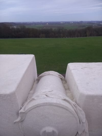 A close-up shows a weathered, stone architectural detail, possibly part of a monument or building. It features a cylindrical element decorated with sculpted leaves, resting on a square block. Beyond, a landscape extends into the distance with a vast green field bordered by a line of trees. A hazy sky tops the horizon, suggesting either early morning or late evening. The angle of the shot gives the impression of looking down from a high vantage point.