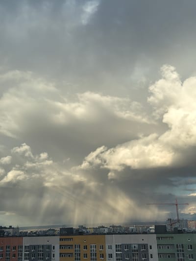 The image, correctly oriented, displays a dramatic sky above an urban landscape, likely captured during sunrise or sunset due to the striking light. The sky is dominated by thick, varied clouds; some are brilliantly illuminated with warm, golden-white light where the sun breaks through, while others appear darker and more grey. A notable detail is the presence of strong, visible sunbeams (crepuscular rays) fanning downwards from behind the clouds on the right side of the frame, creating an ethereal effect.

Along the right side of the image, a row of modern, multi-story apartment buildings forms the cityscape. These buildings feature contemporary designs with varied facades, including panels in light green, grey, yellow, and a reddish-brown hue at the bottom. Numerous windows and some external air conditioning units are visible on their exterior walls. Extending above these buildings and reaching into the clouds is a tall, red construction crane, indicating ongoing development or recent construction in the area.

Beyond the immediate structures, a sprawling urban or suburban landscape stretches into the distance, appearing somewhat hazy under the atmospheric conditions. No people are visible, nor is there any discernible text within the image. The scene conveys a sense of dynamic natural light interacting with a developing urban environment, characteristic of new residential areas like Ankudinovka, Russia.
