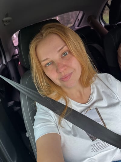 A young woman with blonde hair and freckles is sitting in the driver's seat of a car, looking directly at the camera. She is wearing a white t-shirt and a seatbelt is across her chest. The interior of the car is visible, including the headrest and part of the dashboard. The background is blurred, but appears to be trees and foliage, suggesting an outdoor setting, possibly on a road in or near Novyy Nekouz, Russia.