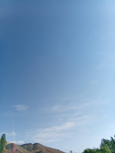 The image shows a wide expanse of a clear blue sky with scattered wispy clouds. In the lower portion of the frame, the top of a mountain range is visible, with earthy tones of brown and tan. Green trees are present along the bottom edge of the image, some appearing as tall, slender deciduous trees and others as denser foliage. A watermark in the lower left corner reads "Galaxy A06". The scene suggests an outdoor natural environment, likely in a rural or mountainous area. Based on the context, this image is situated in the city of Kojo-Aryk, Kyrgyzstan.