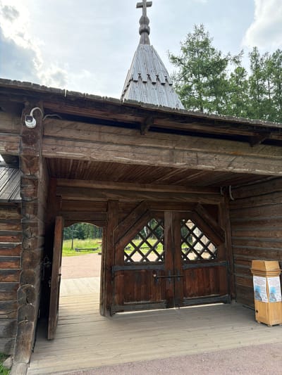 The image depicts an exterior view of a wooden structure in Nevskii Parkleskhoz, Russia, likely a church or an entrance to a religious site. The structure features rustic, log-cabin-style construction with a prominent wooden gate. The gate is divided into two large wooden doors, each adorned with a diamond-shaped lattice pattern. Heavy metal hinges and latches are visible, adding to the historical aesthetic. To the left of the gate, one of the doors is ajar, revealing a glimpse of a pathway and greenery beyond.

Above the entrance, a wooden roof extends outwards, supported by thick logs. Mounted on the underside of this roof is a white security camera with a wire running down a wooden beam. Further up, a multi-tiered wooden roof culminates in a spire topped with a cross. The surrounding environment is characterized by lush green trees, suggesting a park or forest setting. In the foreground, a wooden deck leads to the entrance. To the right of the gate, a wooden information kiosk stands, displaying posters with images of buildings and text in Cyrillic script. The sky above is partly cloudy, with sunlight casting shadows. The overall impression is one of a traditional, well-preserved architectural site nestled within a natural landscape.
