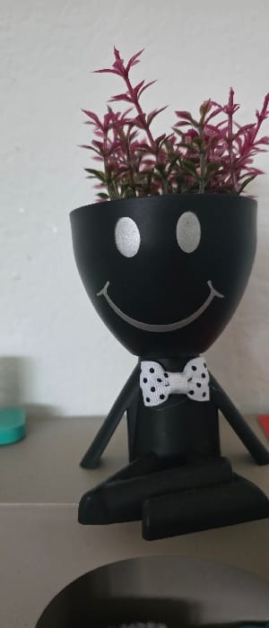 A black, anthropomorphic plant pot, approximately 15-20 cm in height, is depicted in a seated position on a light beige surface. The pot's main body is inverted egg-shaped, featuring a silver-painted smiley face with two oval eyes and a curved mouth. A small, white fabric bow tie with black polka dots is affixed to its neck area. The pot contains artificial-looking plants with slender, reddish-purple leaves. Two black, rounded leg-like protrusions extend forward from the base, resting on the supporting surface. Thin, black arm-like protrusions extend laterally. A plain, light-colored wall serves as the background. Partially visible objects on the supporting surface include a teal-colored circular item to the left and a red item to the right. A dark recess is present below the supporting surface, where the partial word "JADEN" is discernable on an object. The scene is located in Giengen an der Brenz, Germany.