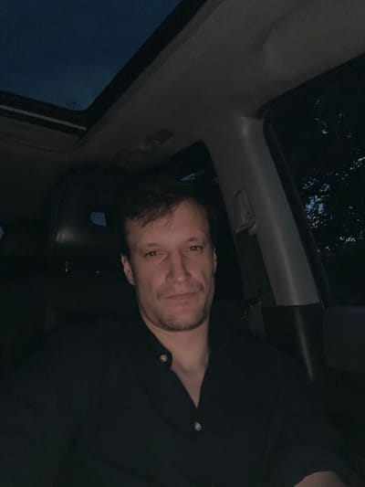 A man is sitting in the driver's seat of a car at night. He is wearing a black collared shirt. The car's sunroof is open, revealing a dark blue sky. Trees are visible through the passenger side window. The interior of the car is dark, with only some ambient light illuminating the scene. It is not possible to determine the specific location in the city of Makiv, Ukraine, from the image.