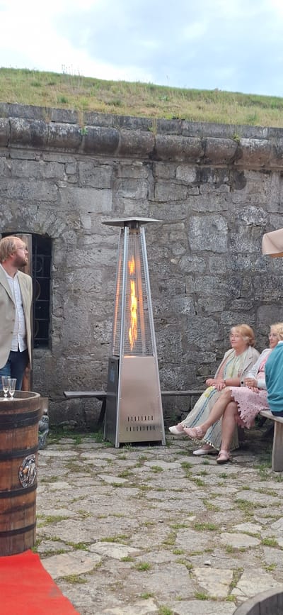 The image depicts an outdoor scene in the city of Kuressaare, Estonia, likely at Kuressaare Castle. In the foreground, a man in a light-colored blazer and white shirt stands to the left, looking towards the left. Next to him is a wooden barrel adorned with a circular medallion. Two champagne flutes are placed on top of the barrel. A tall, pyramid-shaped patio heater with a visible flame is positioned centrally in the mid-ground. Seated on a wooden bench to the right of the heater are two women. The woman on the left wears a patterned dress and a light jacket, while the woman to her right wears a pink dress and a white jacket. The background features a weathered stone wall, characteristic of historical fortifications, with a grassy embankment on top. The ground is paved with uneven, rough-hewn stones interspersed with small tufts of grass. The sky is overcast.