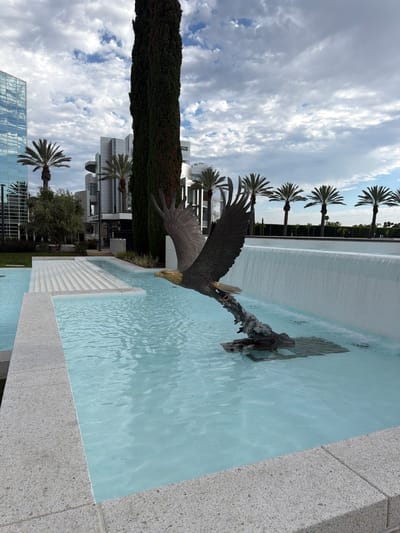 A statue of a bald eagle in mid-flight is prominently featured in a fountain. The eagle statue, made of dark metal, is positioned in the foreground, with its wings spread wide and its beak pointing forward. It appears to be emerging from or diving towards a dark, rocky base submerged in the fountain's water.
The fountain itself is multi-tiered, with the eagle statue in the lower section. The water in this section is a clear, light blue. Behind the eagle, a wide, white waterfall cascades down a smooth, vertical surface, creating a wall of water. The area surrounding the fountain is paved with light-colored stone, with some sections featuring tiered, stepped designs.
In the background, a stark contrast is presented by modern architecture. A tall, slender, dark green tree stands to the left of the waterfall. Further left, a tall, glass-facaded building with a blue reflective exterior dominates the skyline. Other modern, white buildings with curved elements and geometric shapes are visible behind the fountain and palm trees. Several palm trees are spaced out across the midground, their fronds reaching upwards. The sky above is partly cloudy, with patches of blue sky visible between formations of white and gray clouds. The scene appears to be an outdoor public space, likely in a city. This specific location is a public art installation at the Crystal Cathedral in Garden Grove, California, United States of America.