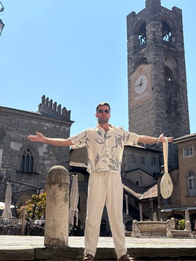 A man stands in a square in front of a tall stone clock tower with a clock face. He is wearing a light-colored short-sleeved button-down shirt with a dark leaf pattern and light-colored drawstring pants. The man is wearing sunglasses and has his arms outstretched to his sides. He is holding a light-colored bag by its strap in his right hand. The square has stone paving, a round stone pillar in the foreground, outdoor seating with umbrellas, and a fountain with lion statues in the background. The architecture visible suggests an Italian setting, possibly in the city of Mozzo, Italy. The sky is clear and blue.