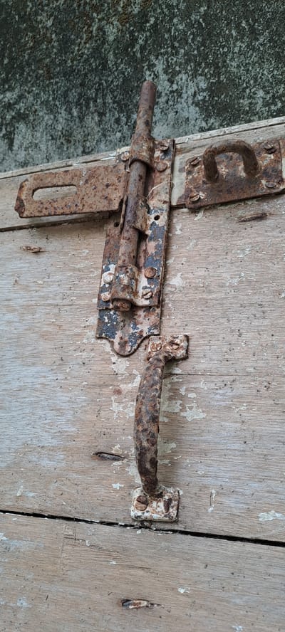 A rusted, old-fashioned slide bolt latch and a matching handle are mounted on a weathered wooden surface, likely part of an outdoor structure or piece of furniture. The latch is a vertical metal rod secured by a bracket with screws, and it slides into a horizontal receiver. The handle is a curved metal piece also attached with screws. The wood shows signs of age and wear, with visible grain and peeling paint. The background appears to be a textured, possibly concrete or stucco wall with some discoloration. The scene is set in Meunasahmeucap, Indonesia, suggesting a tropical or humid environment conducive to the rusting of metal. There are no people or animals present. The focus is on the aged hardware and the wooden surface it is mounted upon.