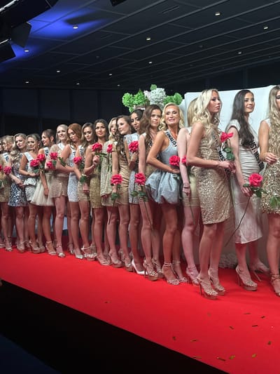 A long row of beauty pageant contestants stand on a red carpet, each holding a red rose. The women are wearing short, sparkly dresses and high heels. They are lined up with their legs visible, most wearing sashes with "FINALIST" or a variation thereof printed on them. At least one has a bouquet of greenery behind her head. The location is likely a stage in Skovshoved, Denmark, judging by the type of event and known beauty contests held there. The background includes a wall with a blue section and a ceiling with square lighting fixtures. Gold confetti is scattered on the red carpet. The contestants are facing the viewer.