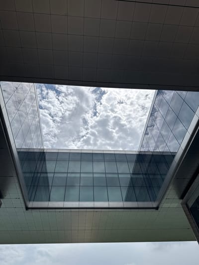 The image shows a perspective looking upwards through a modern architectural structure, likely a sky bridge or atrium, in Spring, Texas. The structure features large glass panels that reflect the sky and surrounding buildings, creating a geometric pattern. The sky is filled with white, puffy clouds against a blue sky. The underside of the structure is covered in a grid of square panels. There are no people or animals visible in the image. The scene conveys a sense of urban architecture and a bright, cloudy day.