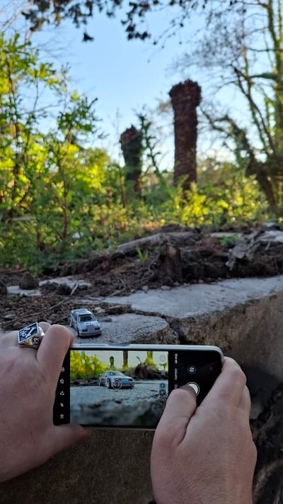 A person is holding a smartphone taking a photo of a toy car in an outdoor environment. The backdrop of the shot includes trees, greenery, and blue sky with a tree in the background. A concrete structure also provides a backdrop to the cars.
The location is not identifiable as Podbadac, Montenegro.