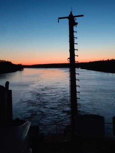 The image presents a view from what appears to be the deck of a vessel, with a body of water in the foreground reflecting the ambient light of the sky. The sky transitions from a deep blue overhead to a reddish-orange near the horizon, indicating either sunrise or sunset. Silhouetted against the sky are trees or foliage along the shoreline, creating a dark, dense line. A dark, vertical structure with ladder-like appendages is prominent in the right side of the frame, possibly a mast or part of the vessel's rigging. The water shows subtle ripples and disturbances, implying movement or wind. The lower portion of the frame contains dark, indistinct shapes that appear to be parts of the vessel's structure or equipment. There are no people visible in the image. The location is Uzhla, Russia.