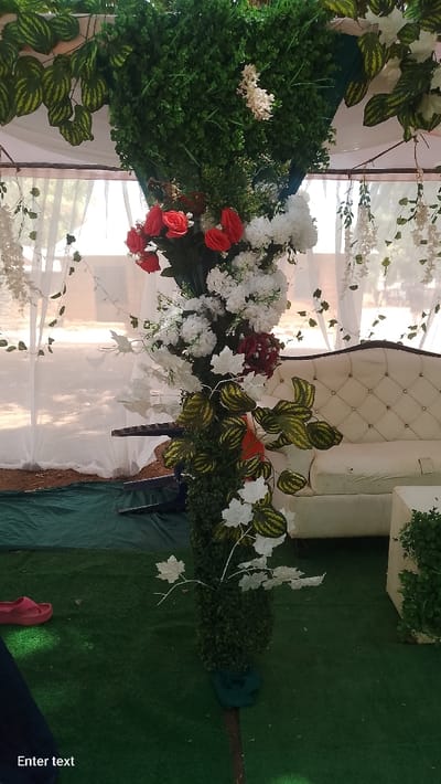 The image displays an event setup in Rinze, Nigeria, characterized by artificial floral and foliage decorations within a semi-enclosed outdoor space. A central vertical column is densely covered with artificial green foliage and adorned with clusters of red roses, white chrysanthemum-like flowers, burgundy blooms, and white and green variegated leaves. Overhead, a section of an artificial green foliage wall is visible, with white hanging floral garlands.
The ground is covered with green artificial turf, beneath which a strip of reddish-brown soil is visible. To the right, a white, tufted, three-seater sofa with a dark wooden frame is positioned on the turf, next to a smaller white cube-shaped object. Sheer white curtains are draped in the background, suggesting a tent or canopy structure. Beyond the curtains, an outdoor environment with trees and open ground under daylight is visible. A pair of red sandals and a portion of dark fabric are present on the artificial turf in the lower-left foreground. No individuals are depicted.