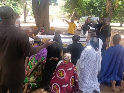 The image shows an outdoor funeral scene in what appears to be Ninzo South, Nigeria. A white casket rests on the ground surrounded by mourners who are kneeling. Several people are standing, some gesturing towards the casket. An ambulance is parked in the background. Many of the mourners are wearing traditional African clothing. One kneeling person is wearing a blue robe. The mourners are gathered under a large tree. The image is taken in daylight.