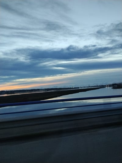 A tranquil scene shows a vast body of water reflecting the cloudy sky above. A strip of land stretches across the horizon, kissed by the faint glow of a setting sun. Silhouetted cranes punctuate the skyline. The foreground is dominated by the blur of a passing structure, with blue guardrails hinting at a bridge or roadway. The overall image conveys a sense of quiet movement and the transition of day into night.