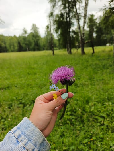 In Medvedevka, Russia, a person's hand, adorned with mint-green nail polish, holds a small bouquet of wildflowers in a lush, green meadow. The dominant flower is a vibrant purple thistle, its spiky petals reaching outwards. It is accompanied by delicate blue forget-me-nots and a single bright yellow buttercup. The background is a soft-focus landscape of a grassy field and a line of birch trees against an overcast sky. The person is wearing a light blue denim jacket.