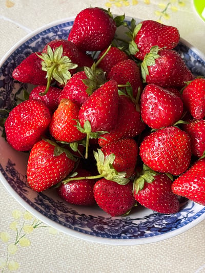 A close-up, overhead shot displays a bowl filled with ripe, red strawberries. The bowl has a white base with a blue, floral pattern around its rim. The strawberries are a vibrant red, some with visible seeds, and green stems and leaves. The bowl sits on a light-colored tablecloth with a subtle pattern of yellow flowers. The scene is well-lit, highlighting the texture and color of the fruit. The image does not depict any people or specific landmarks that would indicate a location in Новый Кинер, Russia.