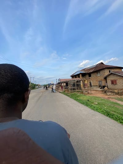 The image depicts a street scene in Igarra, Nigeria, from a low-angle perspective, looking forward. In the immediate foreground, the back of a person's head with dark, short hair and a portion of their left shoulder, clad in a grey-blue garment, is visible on the left. A grey asphalt road occupies the center of the frame, extending into the distance, flanked by dirt verges and green vegetation.

Along the right side of the road, a large, multi-story building constructed from unpainted, rough-hewn concrete blocks or mud bricks features a rusty corrugated metal roof. Several windows and doorways are present, some appearing open or unfinished. Adjacent to this main structure, a smaller, single-story building of similar construction with a rusty metal roof stands. In front of the larger building, a temporary stall-like structure made of wood and corrugated metal is observed. Grass and small plants grow around these buildings.

On the road in the mid-ground, several individuals and motorcycles (likely okadas) are present. One motorcycle is parked on the right side of the road, with a person standing nearby. Further down the road, additional motorcycles and people are discernible, suggesting ongoing activity. Power poles and overhead lines run parallel to the left side of the road, with vegetation and distant structures visible beyond them. The sky is a light blue with scattered, wispy white clouds, indicating daytime conditions.