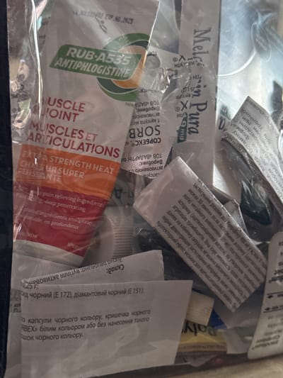 The image shows a close-up view of a collection of medical items, predominantly packaging for various medications. The items are jumbled together, creating a layered and somewhat chaotic composition. Dominating the foreground and left side of the image is a tube or large sachet with text reading "RUB-A535 ANTIPHLOGISTINE" and further down "MUSCLE JOINT MUSCLES ET ARTICULATIONS EXTRA STRENGTH HEAT CHALEUR SUPER PUISSANTE". Below this, smaller text in English indicates "pain relieving ingredients" and "deep penetrating" and in French "ingrédients analgésique en profondeur".
Other visible packaging includes items with Cyrillic text. One prominent label reads "Mel...im Pura", suggesting a foreign origin. There are also smaller blister packs and sachets with various inscriptions, some appearing to be pharmaceutical instructions or ingredient lists. The overall impression is that of an assortment of over-the-counter medications or medical supplies, possibly in a travel bag or medical kit.
The image composition is tightly cropped, focusing on the details of the packaging and text. The lighting is somewhat diffuse, but highlights the reflective surfaces of some of the packaging materials. The background is out of focus, suggesting a shallow depth of field.
The question about the location in Castillo Bajo-conejito, Spain is not relevant to the visual content of the image. The image itself does not contain any visual cues that would place it in a specific geographical location, let alone a city or town.