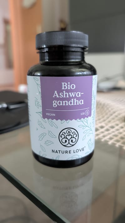 This media file shows a close-up, rotated view of a bottle of "Bio Ashwagandha" supplements. The bottle has a black cap with a ribbed texture for grip. The label is predominantly white with a light purple section containing the text "Bio Ashwagandha" in a vertical, stacked arrangement. Below "Ashwagandha", the word "VEGAN" is printed horizontally. To the right of this text is a circular logo with an ornate design and the number "135" followed by a symbol that appears to be a circle with a line through it, potentially indicating a certification or specific product feature. Further to the right, the text "NATURE LOVE" is printed horizontally, accompanied by small, repeating line-drawn images of leaves and feathers in a light green color. The bottle is placed on a reflective surface, likely glass, which shows a distorted reflection of the bottle and its label. In the background, a blurred, out-of-focus pattern resembling a woven mat or screen is visible. There are no people present. The overall impression is that of a product shot or an unboxing of health supplements. The lighting suggests an indoor setting, possibly a home or studio. The provided location context of Knittelfeld, Austria, does not offer any specific visual cues within the image itself.