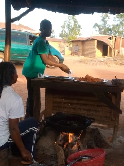 Two women in an outdoor kitchen setting. One stands behind a wooden table, placing food from a plastic bag onto a metal plate; another plate nearby is full of fried food. The second woman sits on a stool in front of a wood-burning stove with a large pot on top. In the background are buildings, a green vehicle, and trees. A red basket sits near the stove.