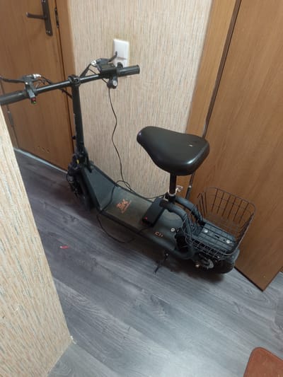 An electric scooter with a seat and a basket is parked indoors, likely in an apartment or hallway, in the city of Hlusk, Belarus. The scooter is dark gray and appears to be in a somewhat dusty condition. It has handlebars with brake levers and a control panel. A black seat is mounted on a post behind the handlebars. In front of the seat, a black wire basket is attached. The scooter is positioned on a gray wood-grain laminate floor. In the background, there is a light-colored, textured wallpaper and a light brown wooden door. A white electrical outlet is visible on the wall. The lighting in the scene is artificial and appears to be from an overhead source.