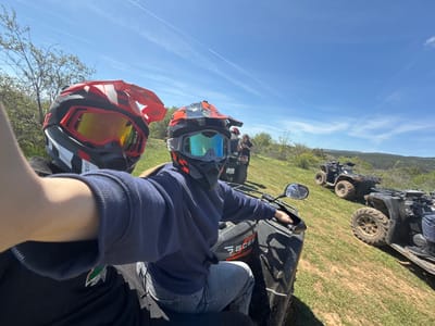 A selfie photograph shows two people in helmets and goggles sitting on all-terrain vehicles (ATVs) in a grassy field. The sky is blue with contrails. In the background, other ATVs and people are visible. The make of the primary ATV is “GOES." The location is outside, but no further detail about Katina, Bulgaria is discernible from the photograph.