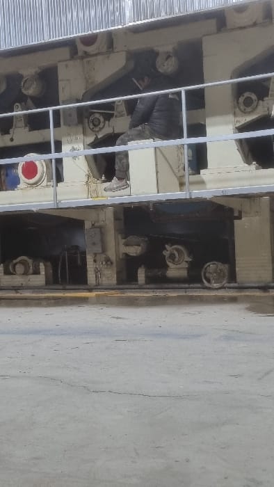 An industrial setting shows large, complex machinery in a predominantly white and gray color scheme. A person is working on the equipment, visible through a metal railing. The machine has various cylinders, gears, and control valves. To the right, a smooth concrete floor or wall is evident, with minor cracks and surface imperfections. The angle of the shot makes the scene appear vertical.