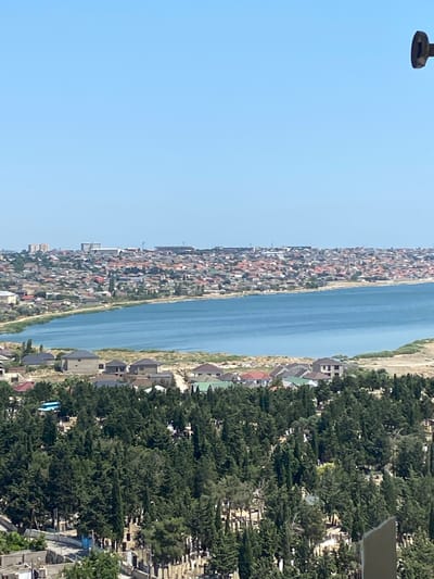 The image captures an elevated panoramic view of a coastal city, likely Bakikhanov, Azerbaijan. The foreground features a densely wooded area with numerous cypress trees, interspersed with what appears to be a cemetery, indicated by visible tombstones and grave markers. Beyond the trees, a body of water, possibly a bay or lake, is prominent. The middle ground displays a stretch of coastline dotted with vegetation and more residences. The background is dominated by a sprawling urban landscape of densely packed houses with red-tiled roofs, extending towards distant buildings and what might be infrastructure like a stadium or industrial structures. The sky is clear and bright blue. A small, dark, metallic object is visible in the upper right corner of the frame.