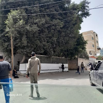 The image shows a street scene in Nuseirat, Palestinian Territories. The shot appears to have been taken from inside a vehicle looking out onto the street, with a partial view of the car visible. On the right side of the frame is a pedestrian walkway made of interlocking tiles with a repeating pattern. The road and the sidewalk are littered with trash and debris.

Several people are visible on the sidewalk. One person is wearing a light-colored coat and dark pants, and appears to be looking at their phone. A woman is sitting near the base of a building with a black hijab. There are other figures further down the street, appearing to be engaged in some kind of market.

The buildings in the background are multi-story and appear to be residential or commercial. A large tree with dense foliage occupies a significant portion of the left side of the image, with many electrical wires hanging from the tree.

A light colored wall forms the backdrop for most of the people. There is a white tarp leaning against the wall.