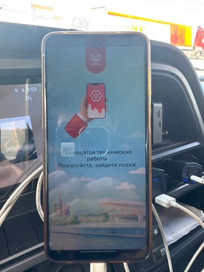 The image shows a smartphone with a light blue screen displaying a message in Russian, indicating that technical work is in progress and asking the user to come back later. The phone is mounted in a car's dashboard. The screen features a logo with a stylized 'M' and the word "АНИМА" below it. A red graphic of a hand holding a phone with a gear icon is also visible. The background of the phone screen shows a picturesque landscape with a stylized depiction of a castle, possibly representing the Kremlin, and clouds.
The surrounding environment appears to be the interior of a vehicle. A second, smaller screen, likely part of the car's infotainment system, is visible to the left of the smartphone. It displays the time "9:53". Various cables and car accessories, including a charging port with a blue light, are present. In the background, through the car's windows, is a blurry view of what appears to be a truck with a white and yellow trailer and a red cab.
Based on the context provided by the logo and the potential Kremlin depiction, this image was likely taken in Il'inskoe-Usovo, Russia, given the Russian language on the screen and the visual cues. The scene is captured in daylight, with bright lighting, suggesting it is daytime. There are no people clearly visible within the frame.