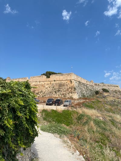 The image shows a view of a large stone fortification under a clear blue sky with scattered white clouds. The fortification appears ancient, constructed of weathered stone blocks. A pathway runs along the base of the fortification, with several vehicles parked on it. These include a silver SUV, a black SUV, and a dark-colored sedan. Lush green vegetation, including dense vines, grows at the bottom of the image, contrasting with the dry, grassy slopes of the fortification's exterior. A metal railing runs along the edge of the pathway, separating it from the natural terrain. A flag, possibly the Greek flag, is visible on a flagpole near the top of the fortification. The scene suggests a historical site, likely a castle or fortress, on a sunny day, possibly in the late morning or early afternoon given the bright sunlight and clear skies. The location appears to be a tourist attraction, given the presence of parked vehicles.