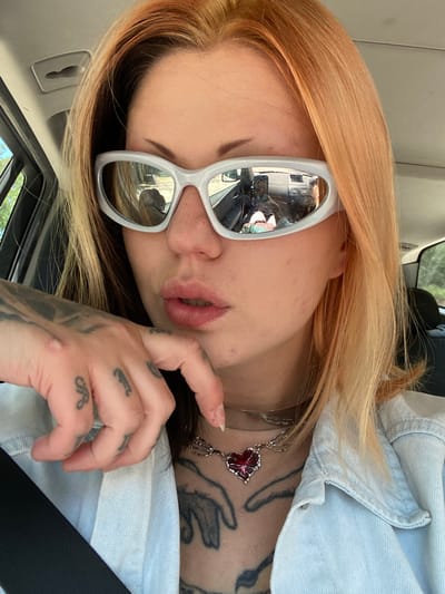 A woman is sitting in a car, facing towards the camera and tilting her head to the left. She is wearing white-rimmed sunglasses, which reflect the inside of the car and exterior scenery. She has light orange hair and is wearing a light blue denim jacket. A black seatbelt is visible across her body. The woman has tattoos on her hand, neck, and chest. She is wearing a silver necklace with a red floral pendant. The location is inside a car, potentially driving through or parked in Kruče - Kërruçi, Montenegro, as suggested by the prompt. The reflection in the sunglasses likely shows the surrounding environment.