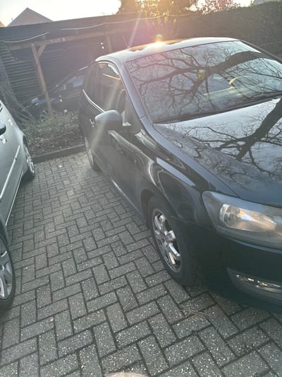 The image displays an outdoor residential parking area in Nettetal, Germany, under strong, low-angle sunlight generating a lens flare in the upper right quadrant. A black, two-door hatchback automobile, consistent with a Volkswagen Polo model, occupies the foreground and midground, parked on grey interlocking concrete pavers laid in a herringbone pattern. The vehicle is equipped with multi-spoke silver alloy wheels. Bare tree branches are reflected prominently across its windshield and hood, with a small yellow decal visible on the inside of the windshield. To the left, a portion of a lighter-colored sedan is visible, also parked on the pavers. In the background, a dark-colored vehicle is parked beneath a wooden carport structure with a slat-style roof. Sparse green shrubbery is present near the carport and between the parked cars. No individuals are present in the scene.