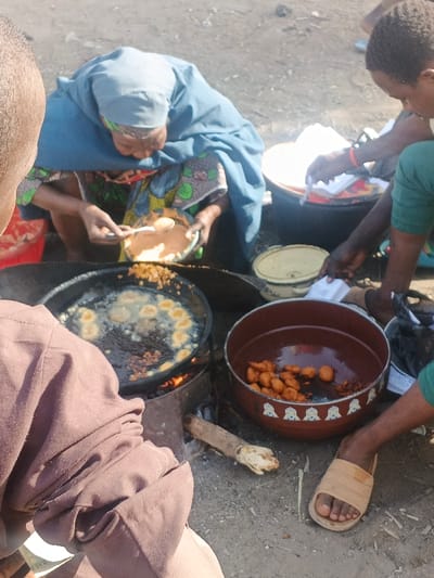 The scene depicts an outdoor food preparation area in Gobirawa, Nigeria. A woman, wearing a blue head covering and a colorful patterned dress, crouches, using a spoon to transfer a light-colored batter from a small bowl into a large black frying pan. The pan is actively heating over an open fire, fueled by visible firewood within a makeshift metal stove, and contains numerous round, golden-brown food items frying in oil. To the right, a large reddish-brown bowl with white decorative patterns holds several already fried, golden-brown items. The lower right arm and hand of another individual, wearing a green garment, are visible near this bowl, handling white paper or packaging. In the foreground, the back and left arm of a third person, dressed in a brown garment, are visible. The ground is unpaved and dusty. A yellow lidded pot and a black bucket containing white items are visible in the background.
