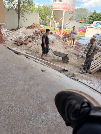 In Příbram, Czechia, a construction site is depicted, featuring a large white cement silo with red lettering. In the foreground, a person's foot and lower leg are visible, wearing a dark sneaker. Further into the scene, two men are present. One man, wearing a dark t-shirt and shorts, pushes a wheelbarrow filled with debris. Another man, dressed in a dark t-shirt and pants, stands with his hands on his hips near stacks of bagged construction material. Debris, including bricks and rubble, is scattered around the area, indicative of demolition or renovation work. A red car is visible in the background, along with fencing and trees. The overall environment suggests an active construction or demolition zone.