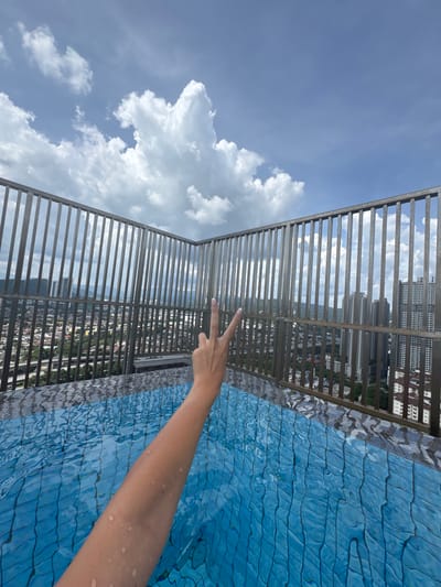 This image captures a vibrant overhead view from a high-rise location, likely a rooftop or elevated pool, in Ampang Jaya Municipal Council, Malaysia. The main subject is a person's wet arm and hand, extending into the frame from the lower right and making a "peace" or "V" sign.
To the right, the bright blue water of a swimming pool is visible, featuring a distinct patterned tile bottom and ripples on its surface, suggesting movement or an infinity edge. A sturdy metal railing, composed of vertical slats, runs across the middle of the frame, separating the pool area from the expansive background.
Beyond the railing, a dense cityscape unfolds under a clear, bright blue sky dotted with fluffy white clouds. Numerous high-rise buildings are visible, indicating an urban environment. The scene is bathed in sunlight, suggesting a clear and sunny daytime. There is no visible text in the image.