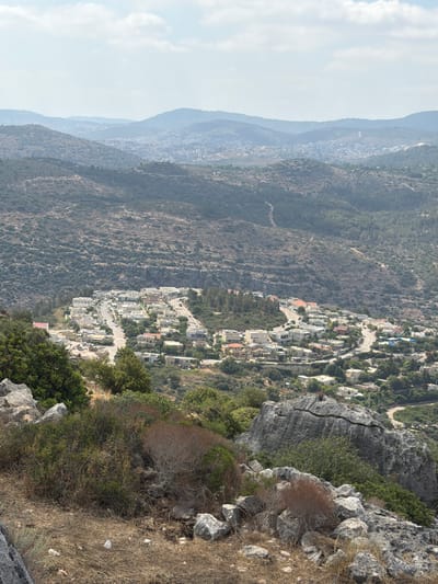 This is an aerial, full shot of a hillside town in the Misgav Regional Council, Israel. The town is nestled within a valley, surrounded by rolling, tree-covered hills. The houses are white and beige with red tiled roofs, clustered together along winding roads. Some of the houses have swimming pools. The foreground of the image is dominated by rocky terrain and dry vegetation, with a large boulder in the lower right corner. The sky is blue with scattered white clouds. The overall impression is one of a peaceful, rural community situated in a beautiful natural landscape.