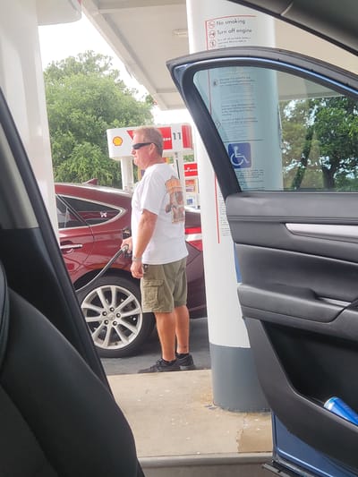 A white man wearing sunglasses, a white t-shirt with a graphic, and olive green cargo shorts is standing at a gas pump, holding the fuel nozzle and fueling a dark red sedan. The car's rear passenger-side door is open, and the fuel door is also open. The man's left foot is resting on the concrete ground. The gas station has white pillars and a covered awning. There are trees in the background. A blue can is visible in the open car door's storage compartment. The scene appears to be in Merritt Island, Florida, United States, based on visible signage from a Shell gas station which are commonly found there. The ground is concrete, with some cracks. The time of day appears to be daytime, likely with overcast skies or shade from the awning.