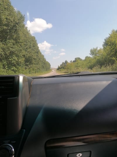 The image is a first-person perspective from inside a vehicle, looking forward through the windshield. The vehicle is on a paved road that curves gently to the right, flanked by dense green trees on both sides. The sky is blue with a few scattered white clouds. In the distance, two white cars are visible on the road. The dashboard of the vehicle is in the foreground, dark and textured, with some metallic trim and a vent visible on the left. A wood-grain trim piece is also visible below the dashboard. There are no people clearly visible in the image. The environment appears to be a rural or semi-rural area with significant tree cover. The location is identified as Severnoe, Kazakhstan.