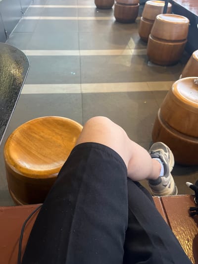 The image shows a person's legs and knees as they sit on a wooden stool. The person is wearing black pants and light-colored athletic shoes. In the background, there are several wooden cylindrical stools and a tiled floor with light-colored stripes. The setting appears to be an indoor public space, possibly a cafe or waiting area. The lighting is natural, suggesting daytime. The perspective is from a seated position, looking down at the person's legs and the surrounding environment. There is no indication of the specific location being Dumai City, Indonesia, or any other city.