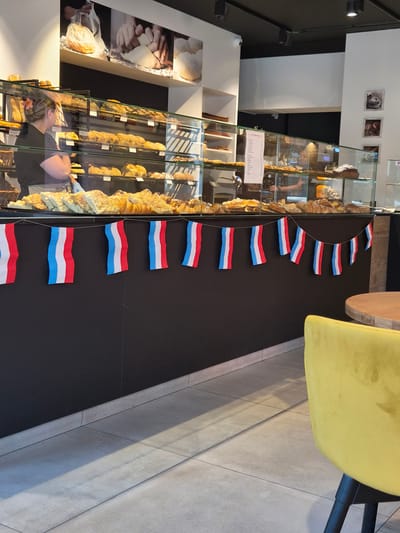A medium shot captures the interior of a bakery, with a focus on the display of baked goods and a decorative string of French flags. In the background, a bakery employee with blonde hair pulled back and a pink flower accessory is visible, wearing a black t-shirt and a white apron. They are standing behind a glass display case filled with various pastries, croissants, and bread. The display case is well-lit, showcasing the products on multiple shelves. Above the display, a large black and white photographic mural depicts hands kneading dough and loaves of bread. A string of small French flags, red, white, and blue, is draped across the front of the counter, adding a festive touch. The lower portion of the image shows a black wall and a light-colored tiled floor. In the foreground, to the right, a corner of a bright yellow upholstered chair is visible, and a portion of a round wooden table. The overall scene suggests a commercial establishment, likely a bakery in Sanem, Luxembourg, given the presence of French flags.