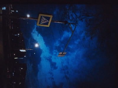 The image shows a street scene at night in Январцев ауылдық округі, Kazakhstan. A pedestrian crossing sign, a triangular sign with a black border and a white background featuring a black silhouette of a pedestrian, is visible. The sign is mounted on a pole. There are also streetlights visible, illuminating the dark sky and trees. The overall impression is of a quiet, dimly lit street.