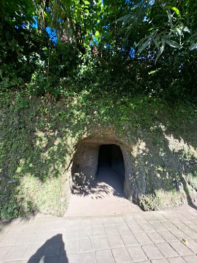 The image shows the entrance to a tunnel carved into a moss-covered hillside. The tunnel entrance is roughly arched and appears to be made of rough-hewn stone. The hillside is covered in lush green vegetation, with sunlight filtering through the leaves of trees above, casting dappled shadows on the ground and the tunnel. The ground in front of the tunnel is paved with interlocking gray stones, and a long shadow of a person is visible on the pavement in the foreground. The tunnel entrance is dark, suggesting depth within. The overall scene is one of a natural, perhaps ancient, structure integrated into a verdant landscape. This entrance is located at Goa Gajah, a significant archaeological site in the region of Banjarangkan, Indonesia.