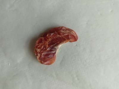 A single dried date sits on a light grey, textured surface. The date is reddish-brown and has an irregular, crescent shape. Its surface appears somewhat wrinkled, with some lighter colored fibrous strands visible. The date is positioned in the lower-middle section of the frame, oriented with its curve facing upwards and to the right. The background is softly lit, creating a subtle shadow that suggests the light source is above and to the left of the object. There is no visible text. The setting appears to be indoors, on a neutral surface.