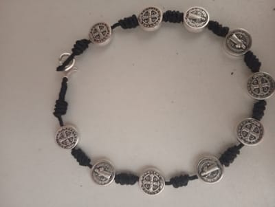 A close-up, elevated shot shows a bracelet on a light-colored surface. The bracelet is made of black cord with silver-toned round charms spaced along its length. Each charm features an embossed religious symbol. The bracelet has a clasp closure with a lobster claw clasp.