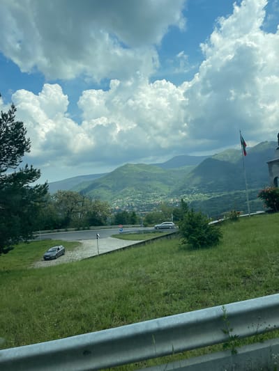The image presents a panoramic view of a mountainous landscape under a partly cloudy sky, observed from an elevated position, specifically in Bunovo, Bulgaria.

In the immediate foreground, a grey metal guardrail runs horizontally across the bottom of the frame, partially obscured by sparse green vegetation. Above the guardrail, a vibrant green grassy slope, dotted with small yellow wildflowers and occasional weeds, ascends towards a paved road and junction area.

The midground features a grey sedan, possibly a station wagon, parked on a dirt and gravel patch beside the road on the left. The road itself is asphalt, curving around and leading into a junction where another silver or light-colored sedan is visible on the right, either stopped or slowly moving. Several road signs are present: a red octagonal "STOP" sign and a circular blue sign with a white arrow (possibly indicating a mandatory direction or roundabout). Lush green trees, including pine trees on the far left and various deciduous trees, frame the road and cover the slopes. To the right of the frame, a tall flagpole displays the national flag of Bulgaria, characterized by horizontal stripes of white, green, and red. Adjacent to the flagpole, a section of a grey concrete or stone building with arched windows is visible, partially adorned with red and orange rose bushes.

The background reveals a series of verdant mountains and hills, densely covered with trees and vegetation, stretching into the distance. A valley is nestled between these mountains, showcasing a small village or town with scattered buildings and infrastructure. The sky above is a mix of bright blue patches and numerous fluffy white cumulus clouds, with some darker grey stratocumulus clouds indicating varied atmospheric conditions.